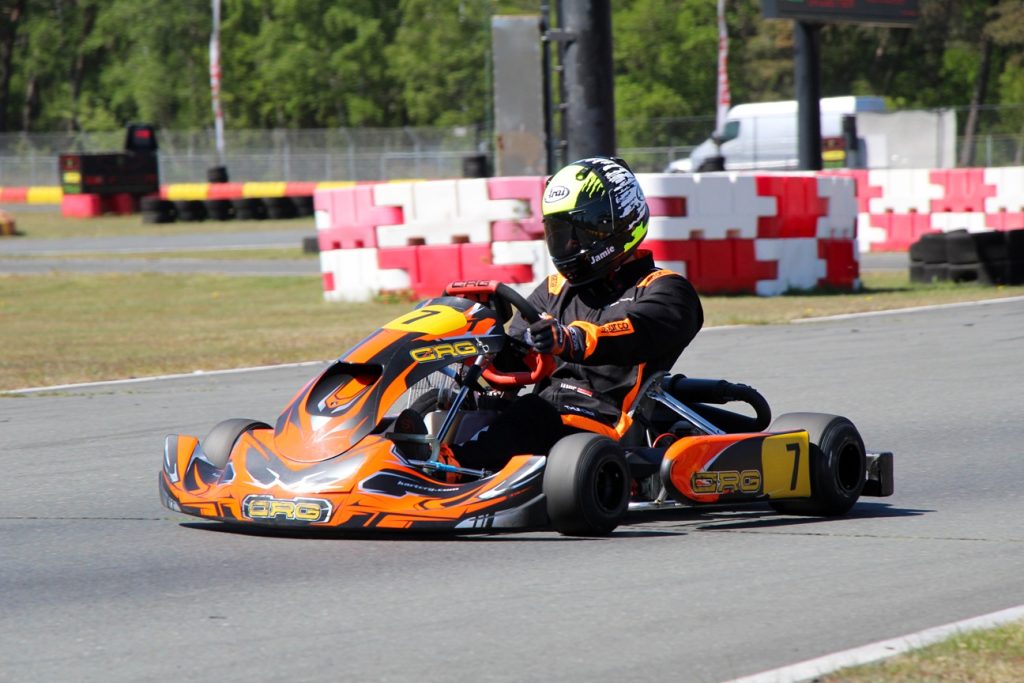 Go-kart racer in black and orange suit driving an orange CRG kart number 7 on a race track with red-white barriers behind.